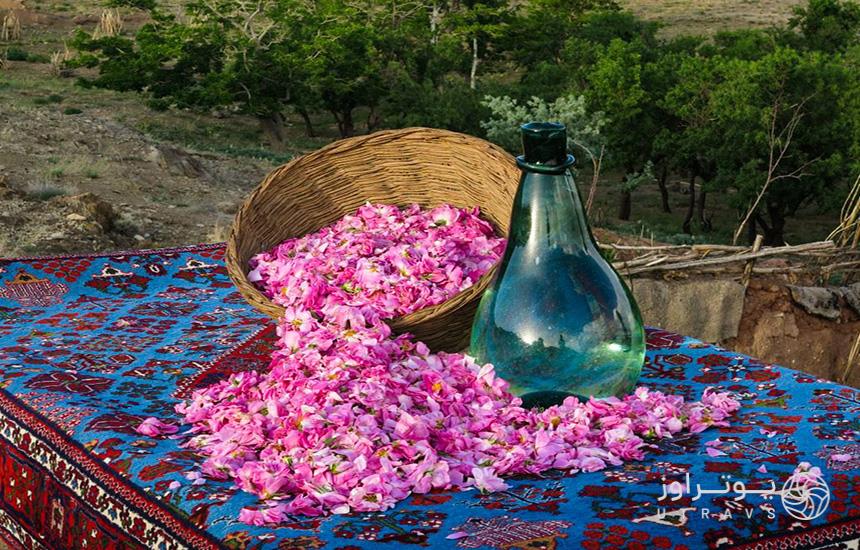 یک سبد حاوی گلبرگ‌های گل محمدی و تنگی سبزرنگ در کنار آن روی قالیچه‌ای به‌رنگ آبی
